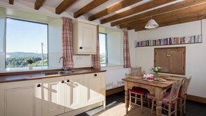 A kitchen with wooden beams, a window with a view, a dining table with chairs, and a bookshelf.