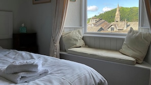Un dormitorio con cama, un asiento junto a la ventana con cojines, un cuadro enmarcado y una mesita con una caja de pañuelos.