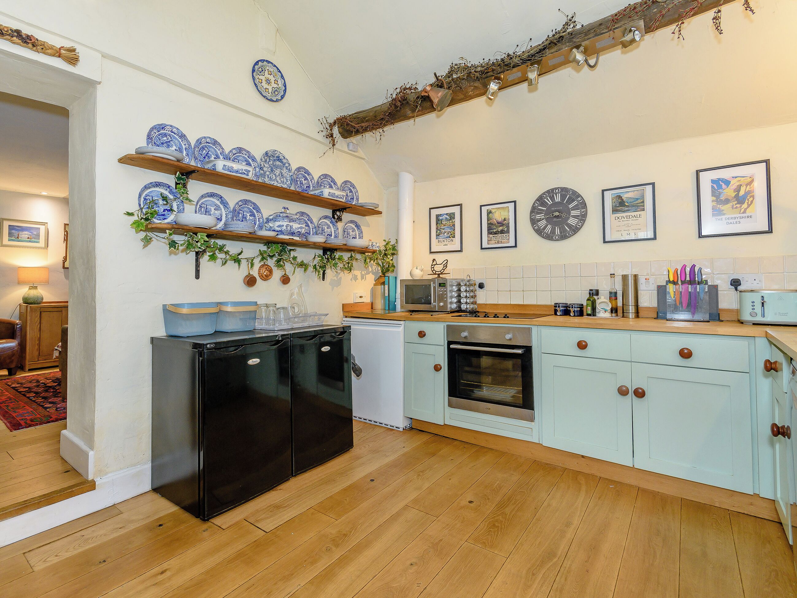 A kitchen with a wooden floor, a black refrigerator, a built-in oven, and a microwave. There are shelves with blue and white dishes and a clock on the wall.