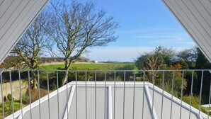 Un balcon doté d’une balustrade en métal, offrant une vue sur un paysage verdoyant, avec des arbres et, au loin, la mer.