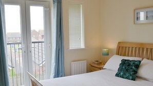 Een slaapkamer met een bed, nachtkastje, lamp, raam met jaloezieën en uitzicht op het omliggende landschap.