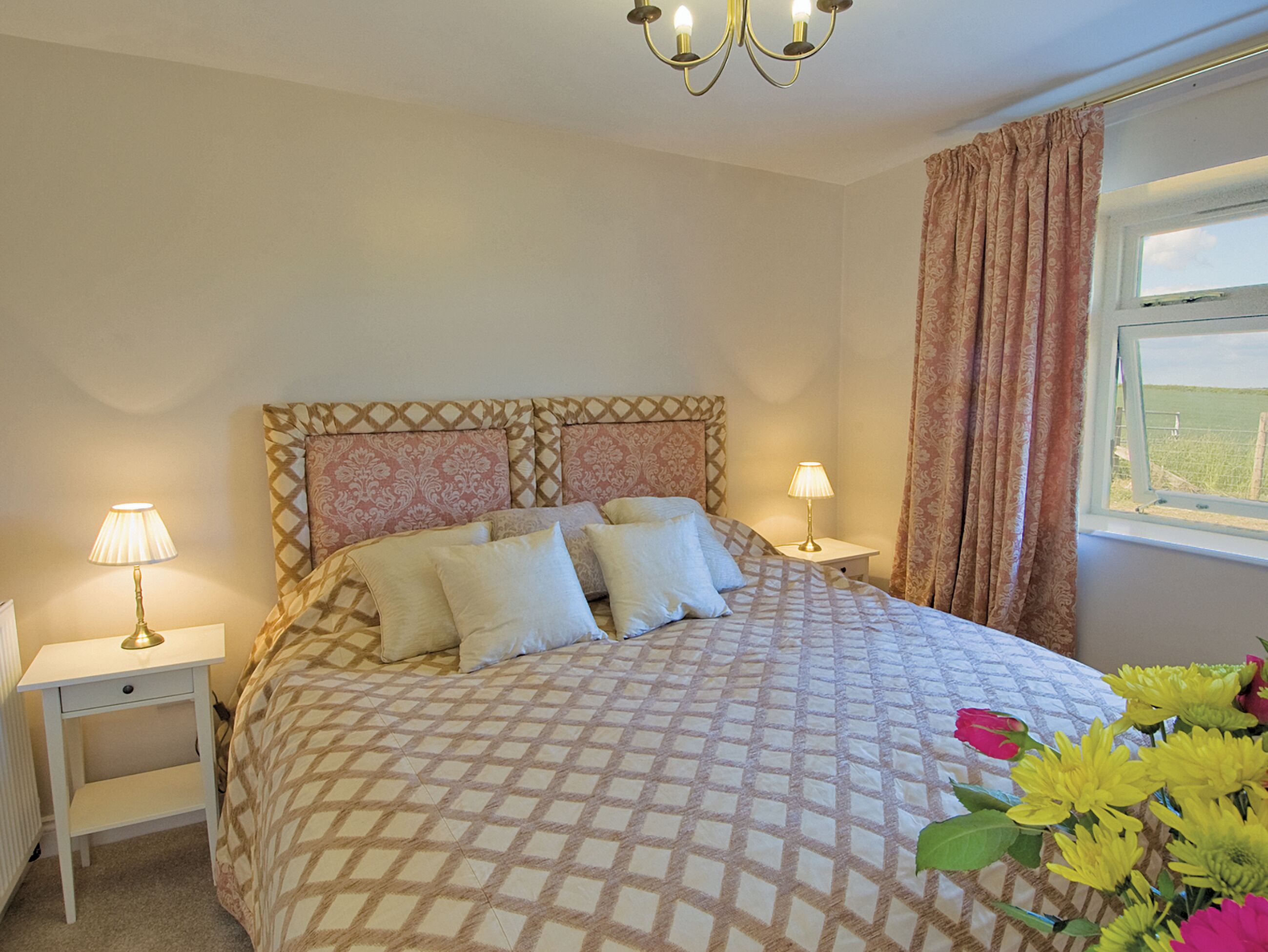 A bedroom with a patterned bedspread, two bedside lamps, a nightstand, and a window with curtains.