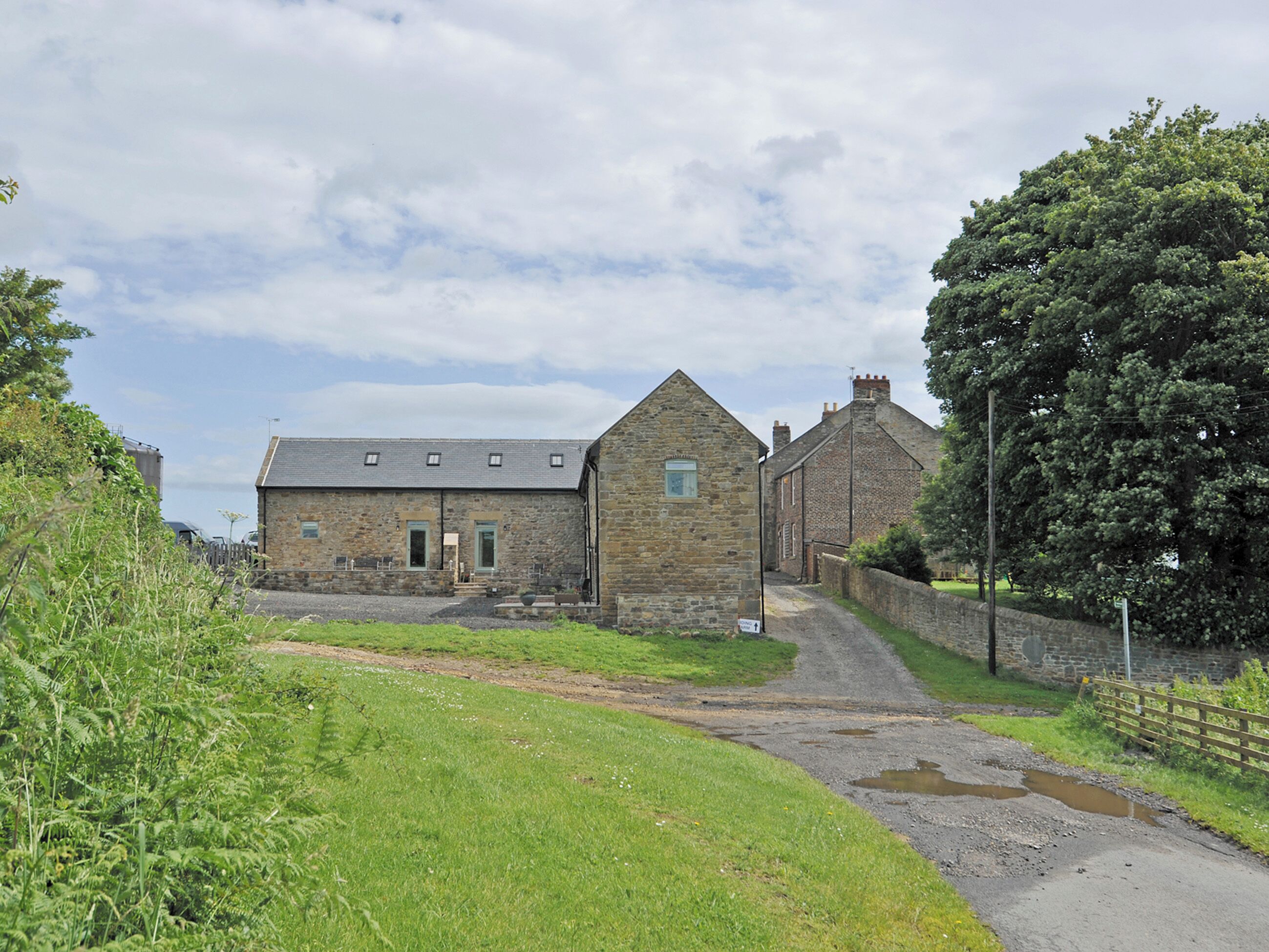 A stone building with a sloped roof, a gravel driveway, and a grassy area with a wooden fence.