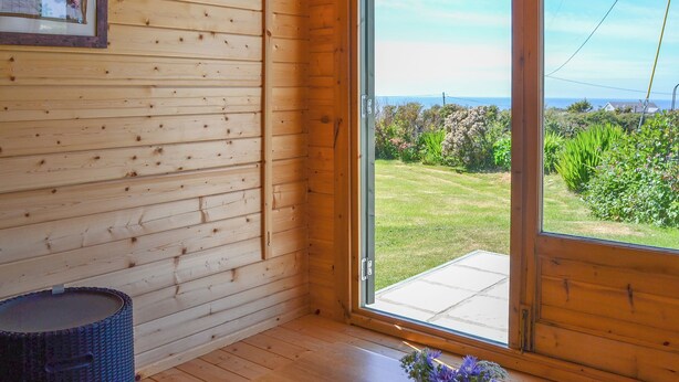 Ein Holzinnenraum mit einer Bank, einem kleinen Tisch mit Blumenvase und einem Fenster mit Blick auf einen Garten und eine ferne Landschaft.
