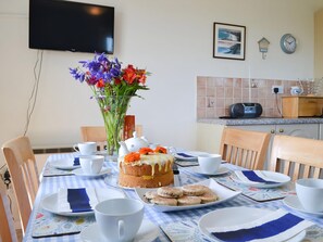 A dining table with a cake, flowers, and biscuits, a TV on the wall, and a kitchen area with a clock and a radio.