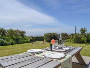 A wooden picnic table set outdoors with plates, a bottle, and a flower. - 3 bedroom accommodation in Church Bay, Anglesey (Church Bay, Anglesey)