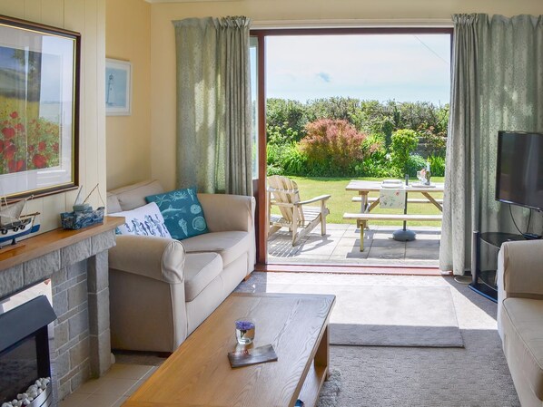 A living room with a sofa, coffee table, fireplace, and a view of a garden through a sliding door. - 3 bedroom accommodation in Church Bay, Anglesey (Church Bay, Anglesey)