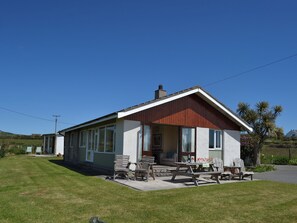A single-story house with a front porch, a dining area with wooden furniture, and a clear blue sky. - 3 bedroom accommodation in Church Bay, Anglesey (Church Bay, Anglesey)