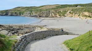 Ein Küstenstrand mit einer Steinmauer, grünen Hügeln und blauem Himmel.
