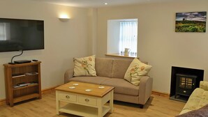 A living room with a flat-screen TV, a wooden coffee table, a beige sofa, a fireplace, and a bookshelf. - 3 bedroom accommodation in Llaneilian, near Amlwch (Llaneilian, near Amlwch)