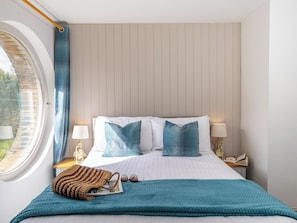 Una cama bien hecha con sábanas blancas y almohadas azules, una ventana con vistas a la vegetación, y una mesita de noche con una lámpara y un libro.