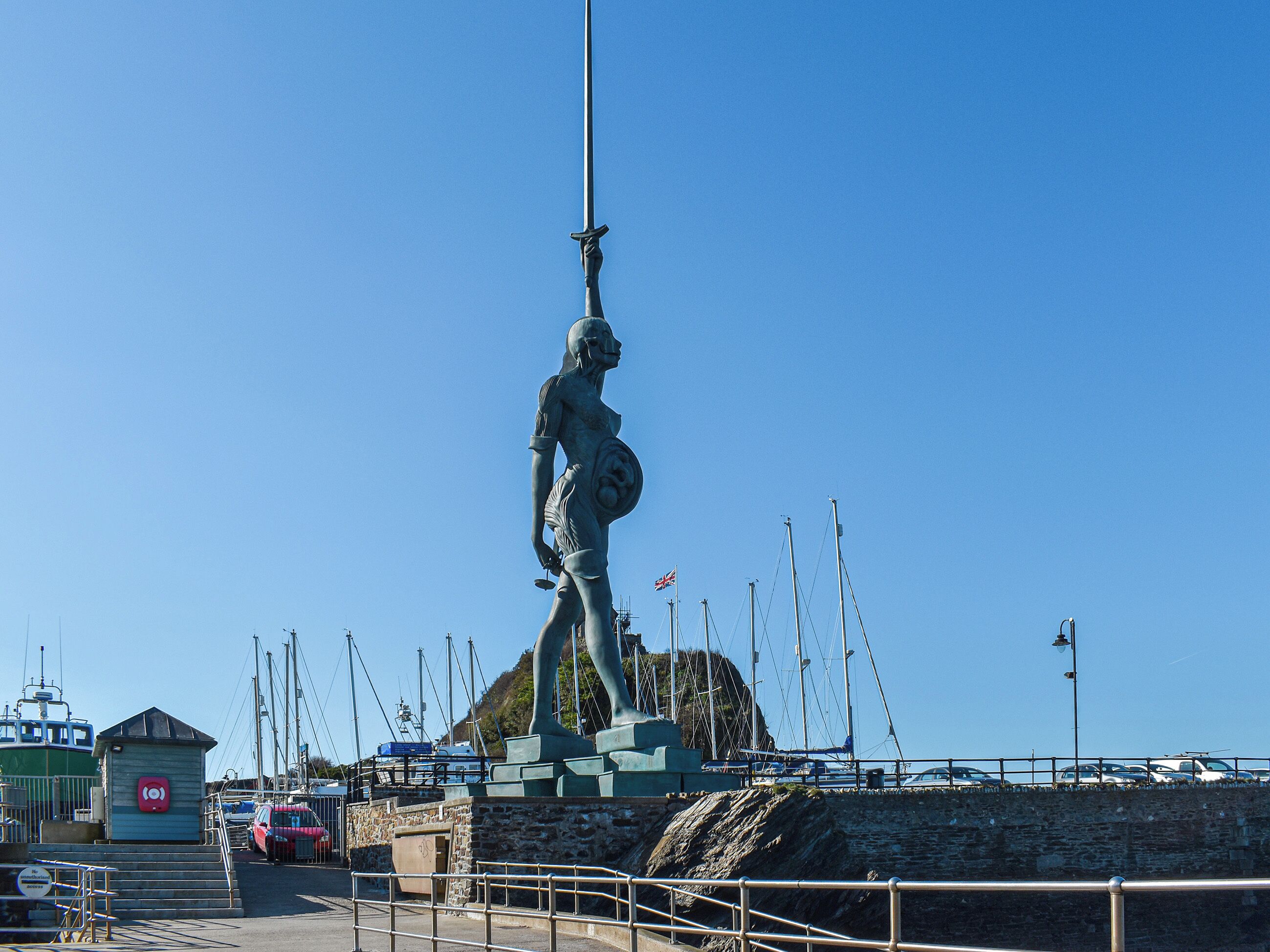 A statue of a woman holding a shield and spear, located near a waterfront with boats and a small building.