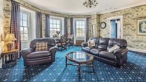 A room with a blue patterned carpet, leather sofas, a glass coffee table, and a chandelier. - 2 bedroom accommodation in Ilfracombe (Ilfracombe)
