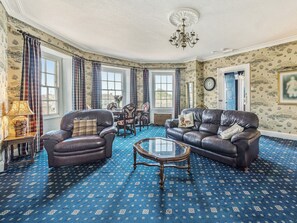 A room with a blue patterned carpet, leather sofas, a glass coffee table, and a chandelier.