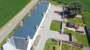 Vue aérienne d’une propriété comprenant un long bâtiment étroit, une cour clôturée et un grand champ ouvert.