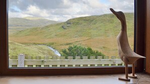 A wooden bird sculpture on a windowsill overlooking a landscape with hills and a stream.