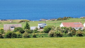 Un paysage côtier avec vue sur la mer, comprenant une maison moderne avec un toit rouge et des panneaux solaires, entourée de verdure et d’un champ herbeux.
