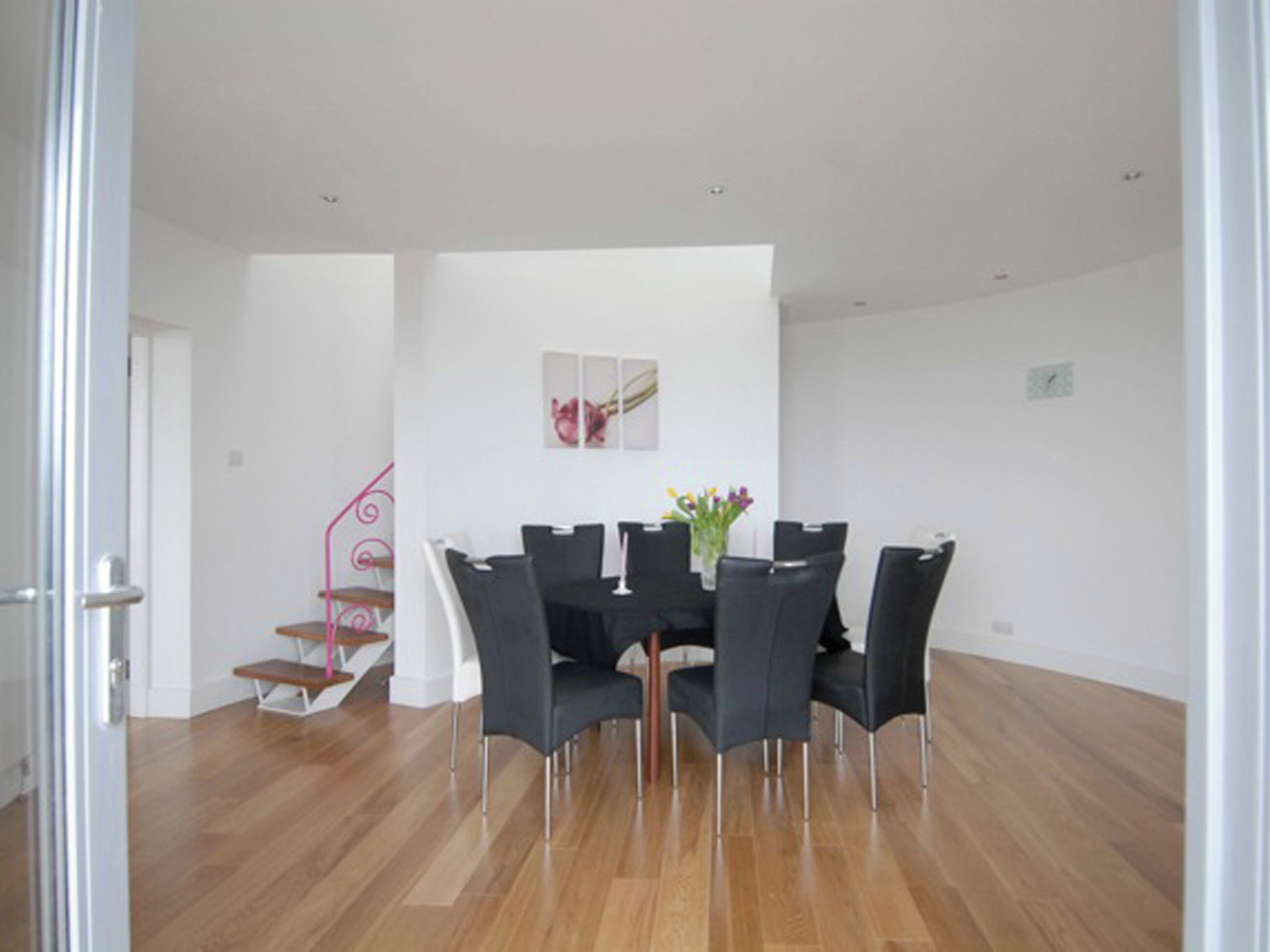 A dining area with a wooden floor, a table with chairs, a vase with flowers, and a staircase with a pink railing.