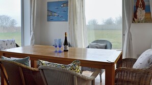 A dining area with a wooden table, wicker chairs, and a view of the countryside through large windows.