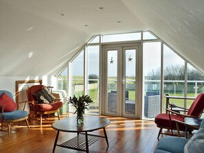 A spacious room with a sloped ceiling, large windows, and a view of the outdoors. There are two chairs and a table with a vase of flowers.