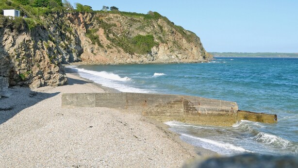 A pebble beach with a rocky cliff and a small white building on top.