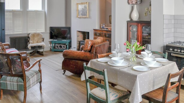 Un comedor con una mesa dispuesta para cuatro, televisor, chimenea y zona de cocina con estufa.