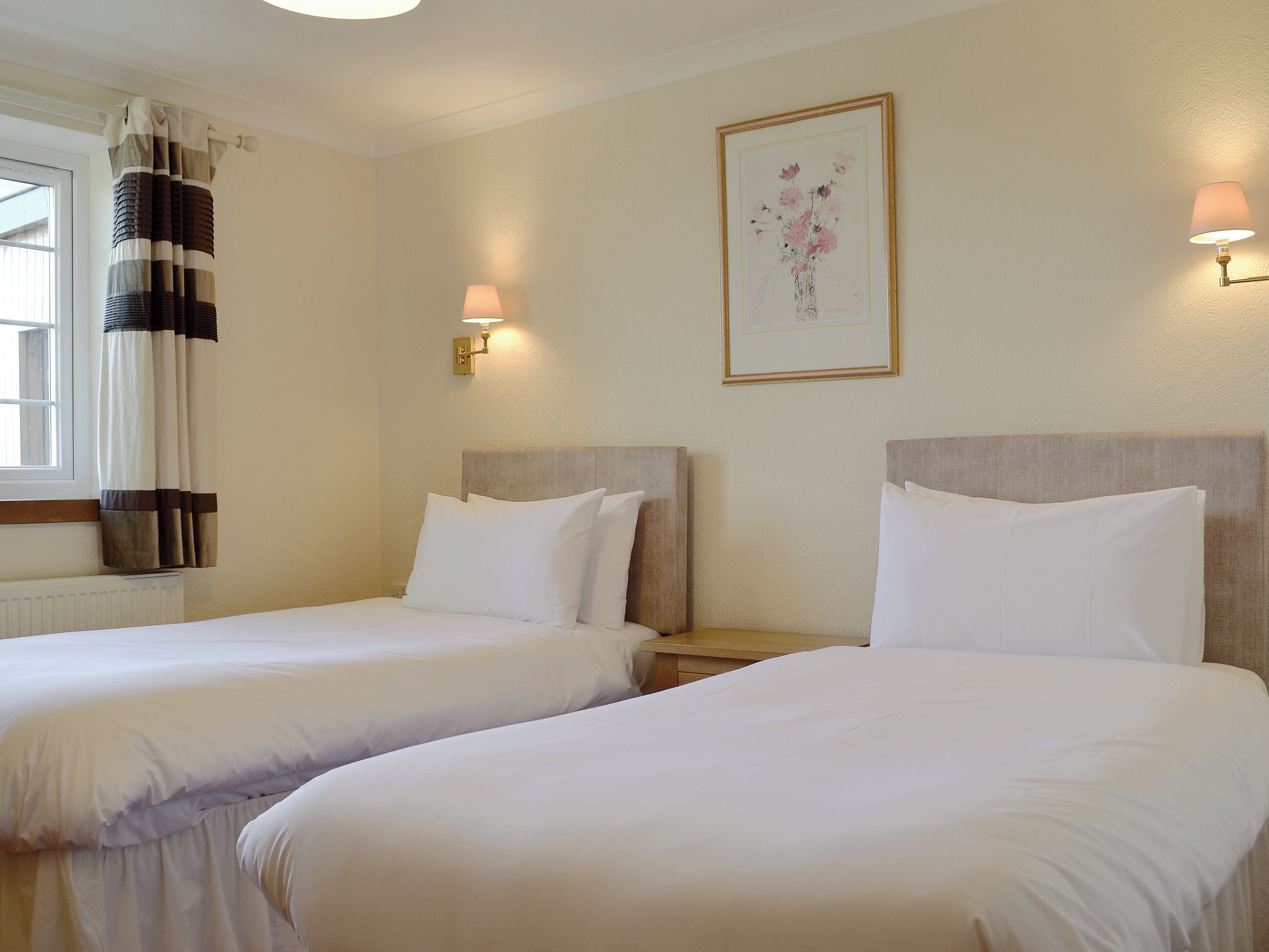 A hotel room with two single beds, a window with curtains, a wall-mounted lamp, and a framed floral artwork.