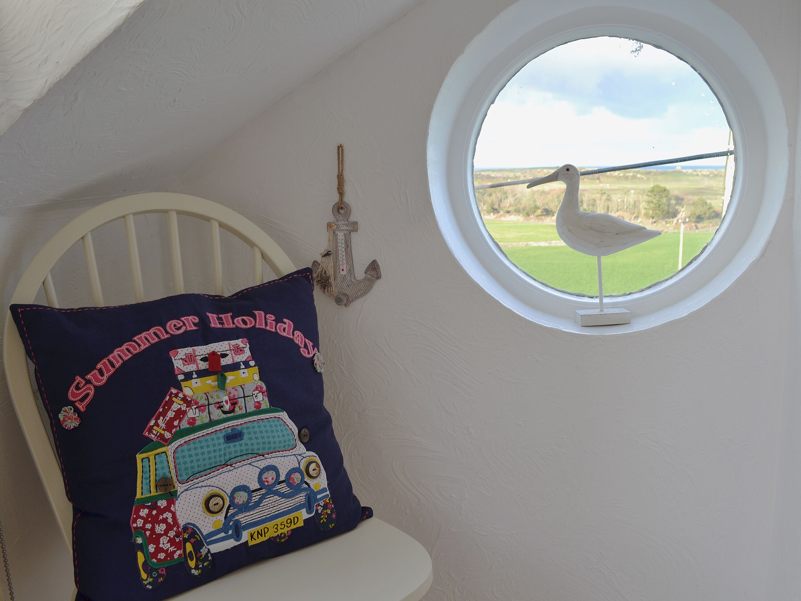 A bedroom with a window, a pillow with a summer holiday design, and a wall-mounted anchor decoration.