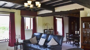 A living room with a black leather sofa, a wooden coffee table, and a chandelier. There is a window with red curtains and a view of greenery outside. - Kiln Green Farmhouse (Near Milnthorpe)