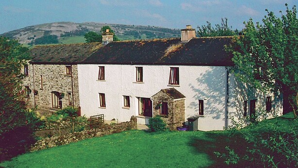 A white building with multiple windows and a stone structure attached to it, surrounded by greenery and hills in the background. - Kiln Green Farmhouse (Near Milnthorpe)