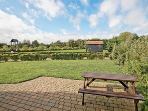 A picnic area with a brick-paved surface, a wooden picnic table, and a playground structure in the background.