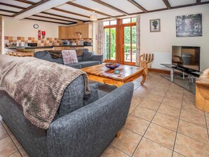Una sala de estar con un sofá, una mesa de centro y un televisor. Se observa una cocina al fondo con armarios de madera y una ventana.