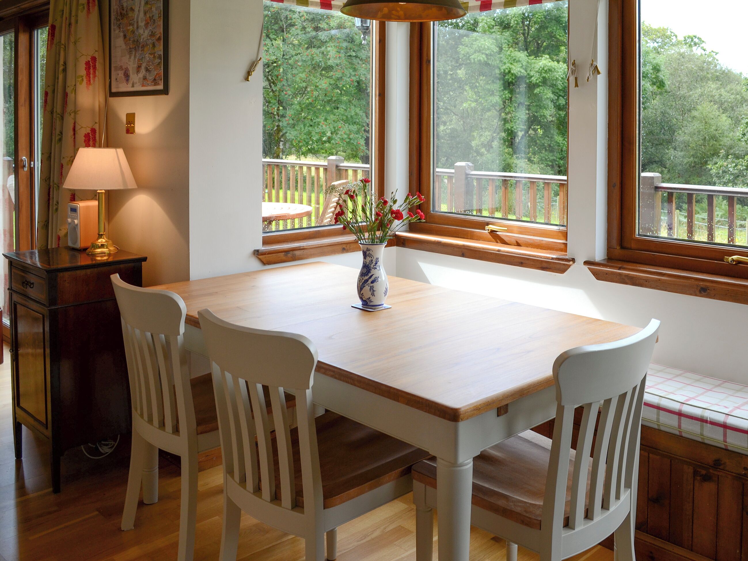 Uma área de refeições com mesa de madeira e cadeiras brancas, um vaso com flores, e vista para um deck de madeira e árvores através de grandes janelas.