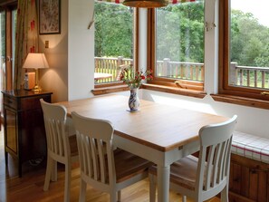 A dining area with a wooden table and white chairs, a vase with flowers, and a view of a wooden deck and trees through large windows.