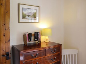 A wooden dresser with three drawers, a small shelf with books, a framed landscape painting, and a bedside lamp. - Allt Beag (Little Streams) (Achintraid, near Lochcarron)