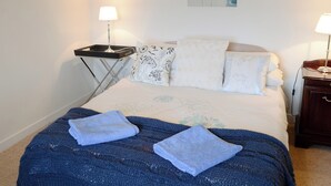 Un lit bien fait, avec une couverture bleue et deux serviettes, une table de chevet avec une lampe, et une œuvre d’art accrochée au mur au-dessus du lit.