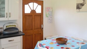 Une cuisine avec des meubles blancs, une porte en bois, une table recouverte d’une nappe à motifs floraux et une radio posée sur le comptoir.