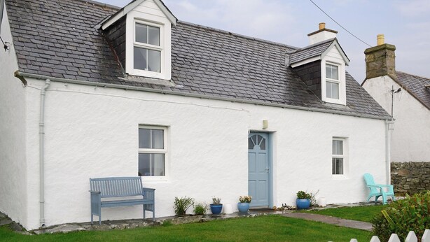 Une petite maison blanche avec un toit recouvert de tuiles grises, une porte bleue et une clôture en bois peinte en blanc.