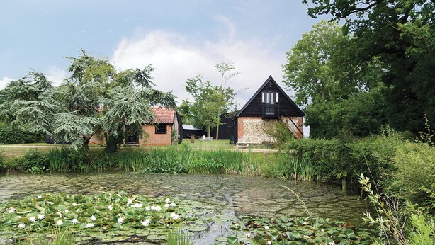 Um edifício rústico com telhado inclinado, rodeado por vegetação exuberante e um lago com nenúfares.