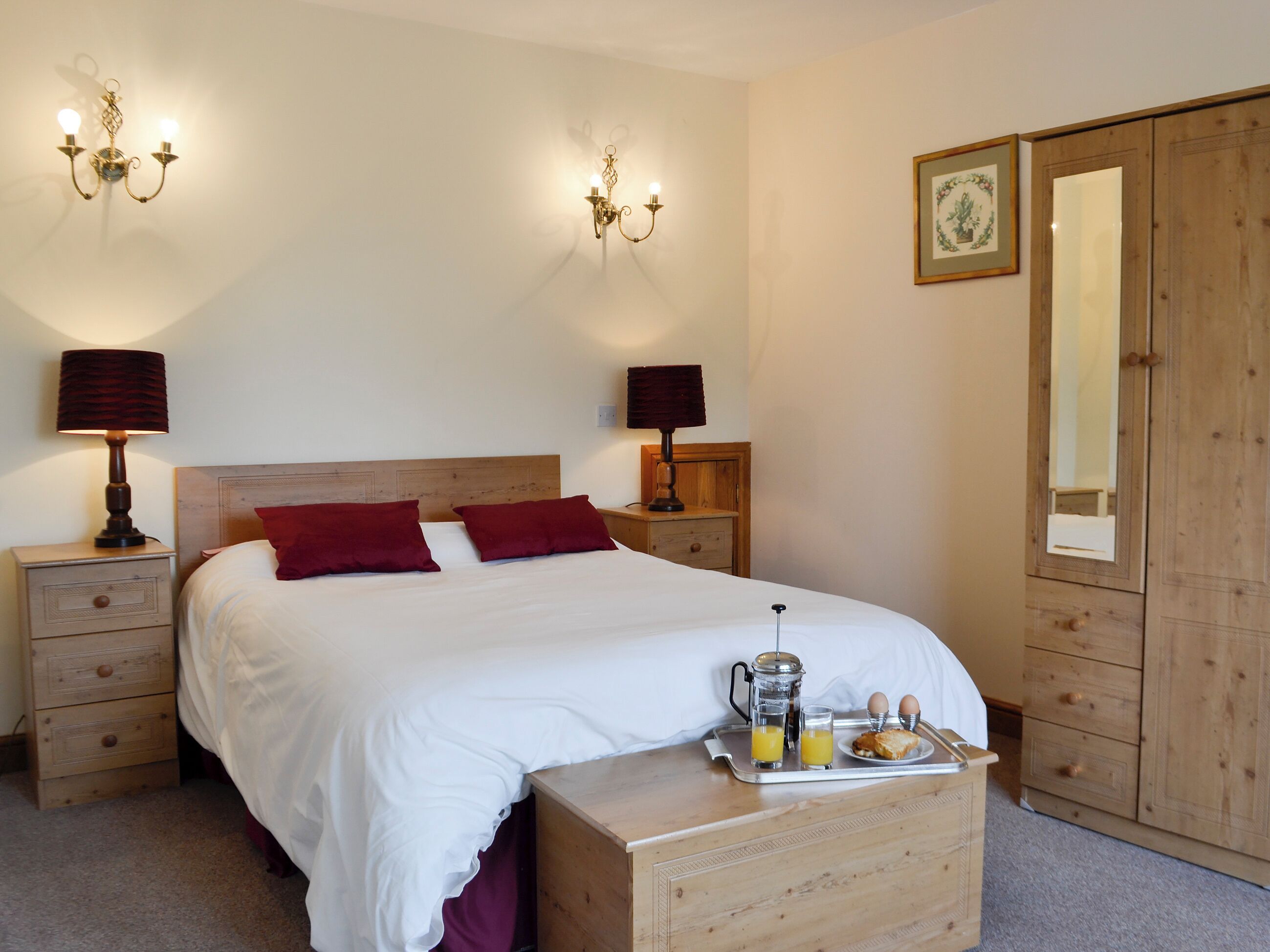 A neatly made bed with two pillows, a bedside table with a lamp, a tray with breakfast items, and a wardrobe with a mirror.