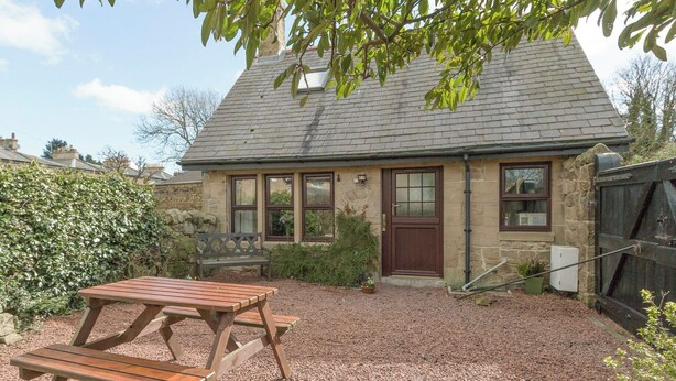A stone cottage with a tiled roof, a wooden bench, and a gravel driveway.