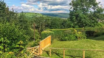 A rural landscape with a wooden gate, lush greenery, and a distant view of hills.
