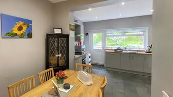 A dining area with a wooden table, chairs, a vase of flowers, a framed picture, and a kitchen in the background.