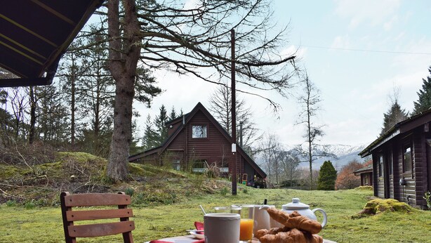 Ein Holztisch mit Frühstücksangeboten, ein Stuhl und Blick auf Blockhäuser und Bäume.