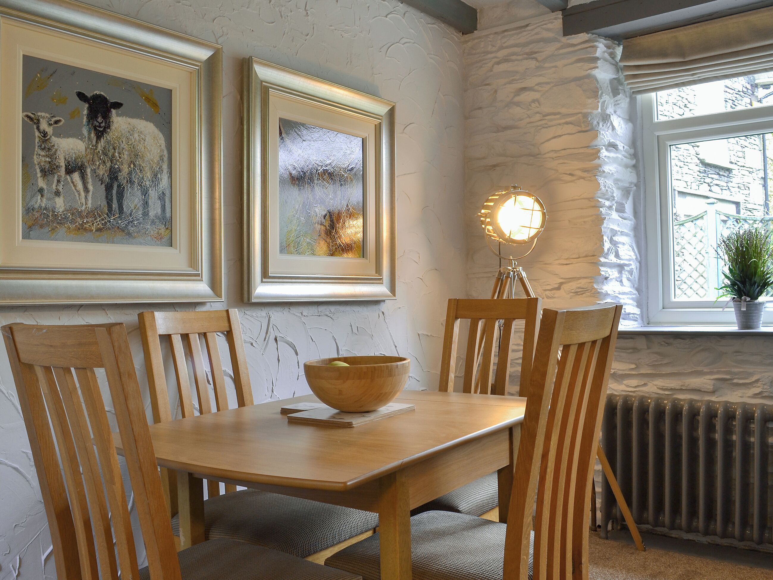 A dining area with a wooden table, chairs, a bowl, framed artwork, a lamp, and a window with a view.