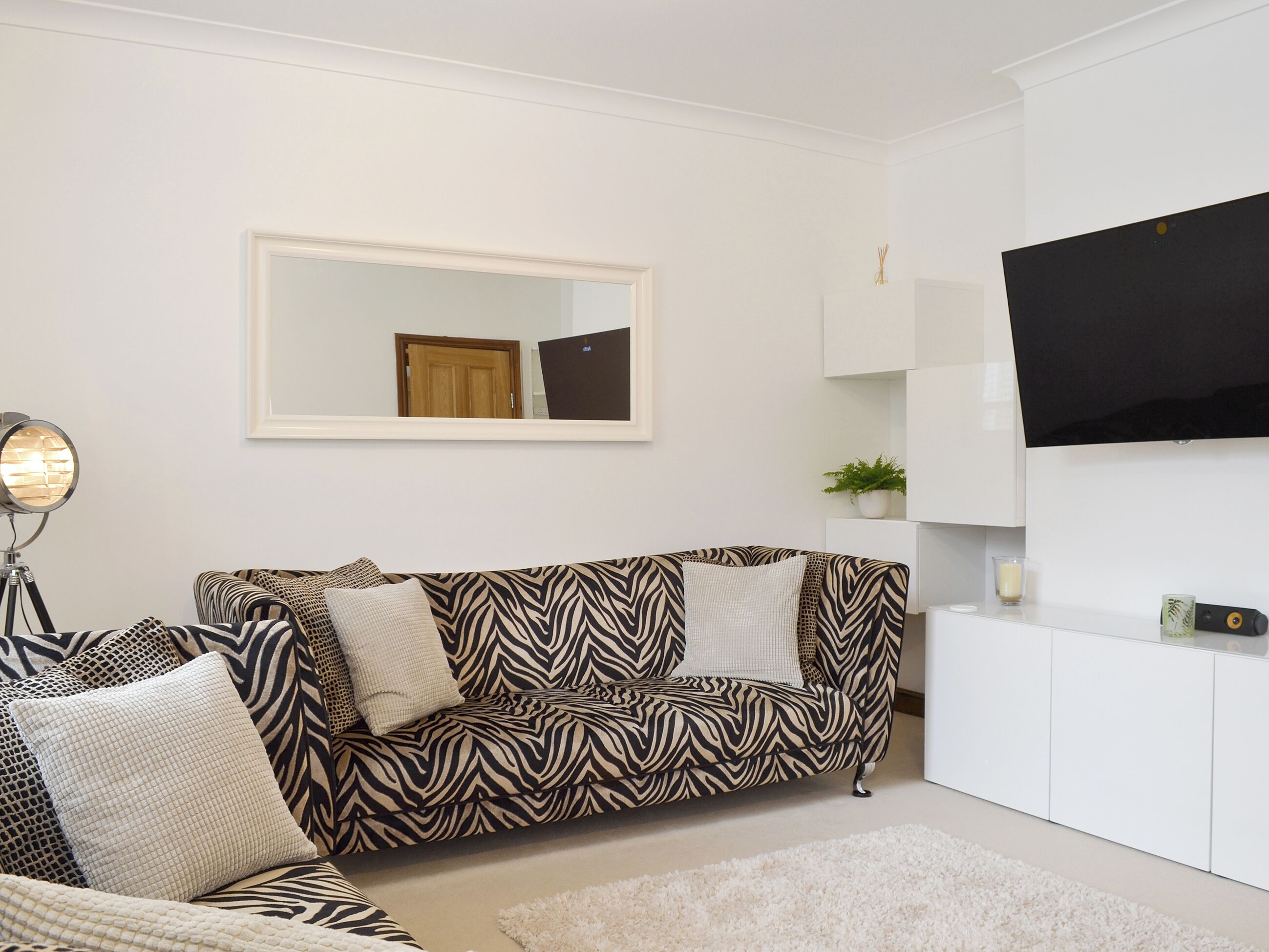 A modern living room with a zebra-patterned sofa, a white armchair, a floor lamp, a large mirror, a flat-screen TV, and a small potted plant.