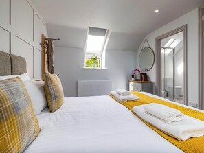 Une chambre d’hôtel moderne avec un grand lit, une petite fenêtre, un radiateur, une table de chevet avec un miroir et une salle de bain visible à travers une porte en verre.