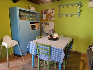 Un comedor con una mesa dispuesta para dos, un armario azul con utensilios de cocina, un perchero para tazas en la pared y un tablón de anuncios con varios avisos.