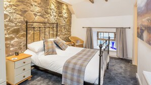 Une chambre avec un mur en pierre, un lit avec une couette à carreaux, une table de chevet, une chaise et une fenêtre avec des rideaux.