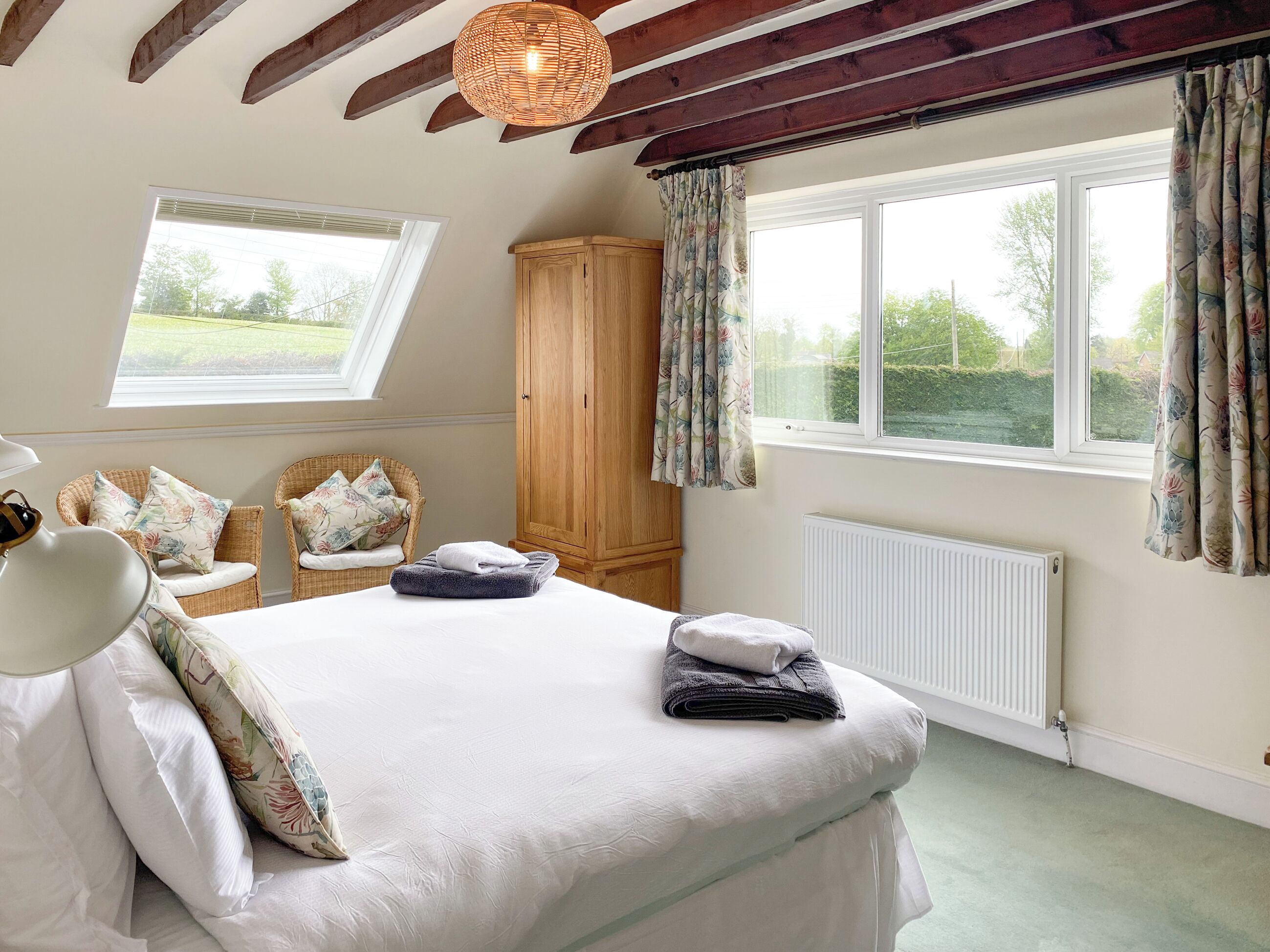 Ein Schlafzimmer mit einem Bett, zwei Korbstühlen, einem Holzschrank, einem Heizkörper und einem Fenster mit Vorhängen.
